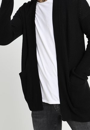 Homme portant un t-shirt blanc, un cardigan en maille foncé avec des poches, et un jean sombre, une main levée près de la tête.