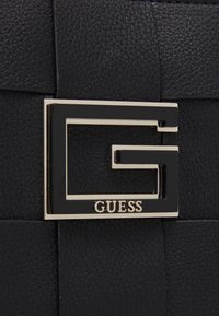 Svart strukturerad läderhandväska med en framträdande svart och guldfärgad "G"-logotyp och "GUESS"-märkning under.