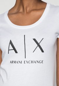 Bílá tričko s logem "A|X ARMANI EXCHANGE" v tučných černých písmech, s výstřihem do lodi a přiléhavým střihem.