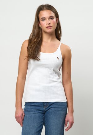 Young woman with long brown hair wearing gold hoop earrings, a white sleeveless top with a small embroidered logo, and blue jeans, standing.