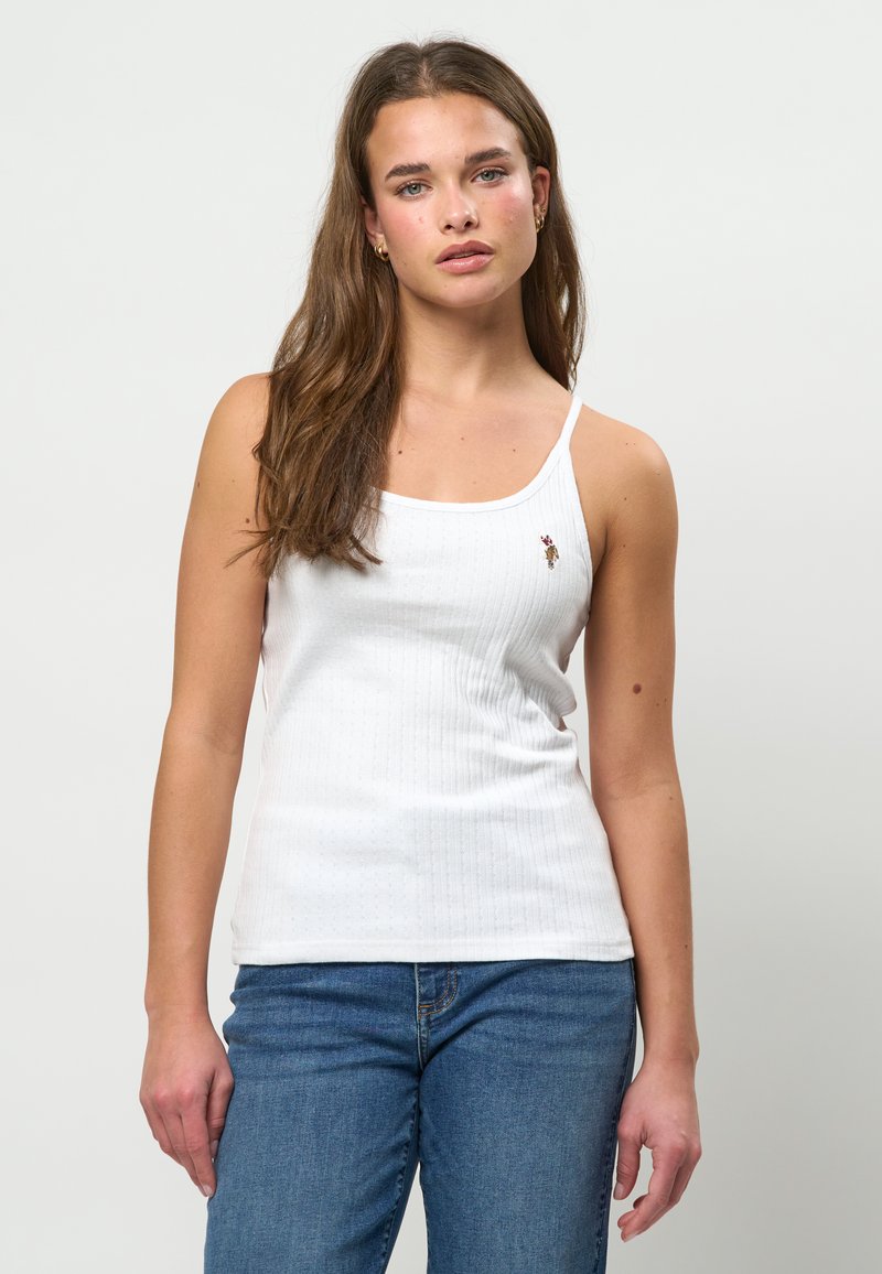 Mlada ženska z dolgimi rjavimi lasmi, ki nosi zlate obročaste uhane, belo brezrokavnik z majhnim vezenim logotipom in modre kavbojke, stoji.