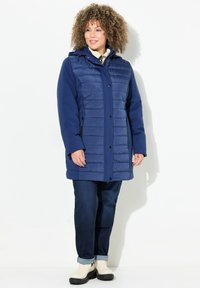 Giacca lunga blu navy con parte frontale trapuntata e maniche lisce. Presenta un cappuccio, bottoni a pressione e tasche laterali con zip. Indossata con jeans e stivali.