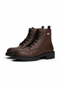 Braune Leder-Stiefeletten mit schwarzen Schnürsenkeln, runder Spitze, dicke schwarze Gummisohle und einem Logopatch an der Seite. Robuste Bauweise und Textur.