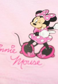 T-shirt rose clair avec un graphique représentant un personnage de dessin animé portant une robe à pois et un nœud, avec des accents noirs et du texte stylisé en dessous.