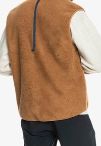 Brun fleeceväst med en texturerad yta, med en marinblå accent vid kragen och utan ärmar. Minimalistisk och funktionell form.