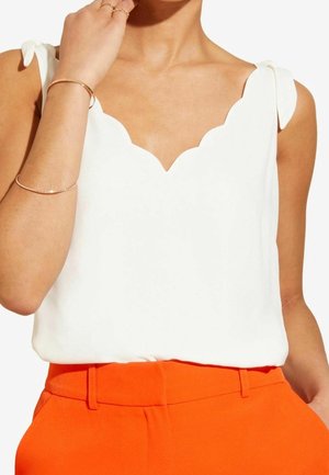 Kvinde iført en ærmeløs hvid top med bølget halsudskæring og bundne skulderstropper, kombineret med orange bukser med høj talje.