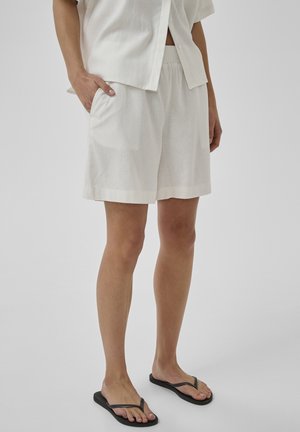 Person iført løse hvide shorts og en matchende kortærmet skjorte med den ene hånd i lommen, sorte flip-flops på fødderne.