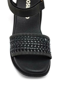 La sandalia negra presenta una banda trenzada en la parte superior, una correa suave y una plantilla texturada con una suela plana para mayor comodidad.