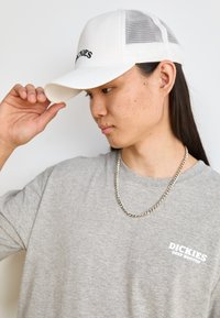 White mesh-back cap with embroidered logo, gray t-shirt with small chest print, and silver chain necklace. Side profile view.