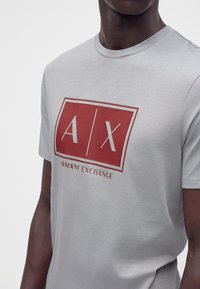Άνδρας που φοράει ανοιχτό γκρι μπλουζάκι Armani Exchange με κόκκινο τετράγωνο λογότυπο και λευκά γράμματα A και X μπροστά.