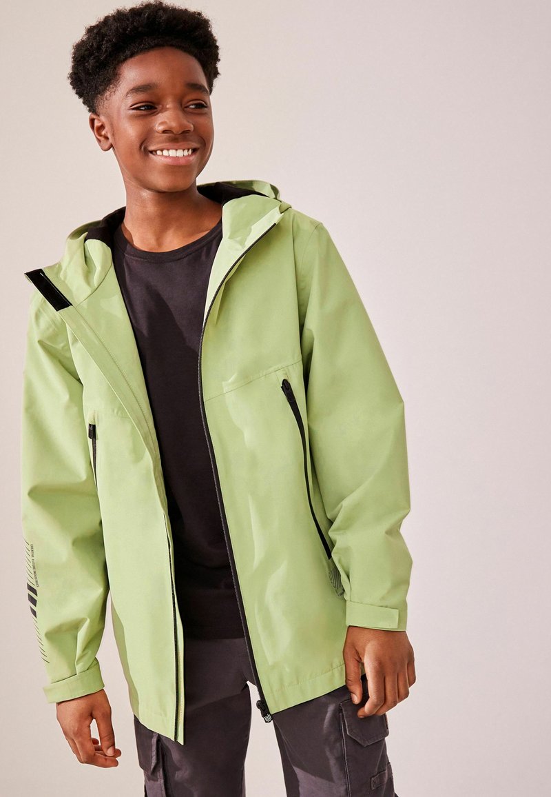 Grüne, wasserdichte Jacke mit Reißverschluss vorne, Kapuze und schwarzen Akzenten. Verfügt über Reißverschlusstaschen und eine glatte Textur. Getragen über einem schwarzen Shirt.