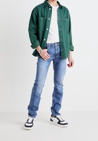 Person klädd i en grön skjorta med knappar över en vit t-shirt, ljusblå jeans, vit och svart sneakers, stående mot en ljus, enfärgad bakgrund.