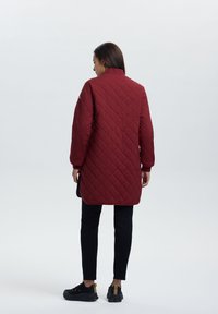 Gevoerde bordeauxrode lange jas met hoge kraag, met een rechte snit en een paar zijzakken, gedragen over zwarte broek en schoenen.