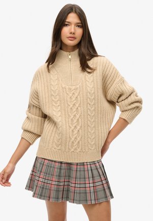 Suéter beige de punto con cuello alto y cremallera. Presenta patrones texturizados de trenzado. Combinado con una falda plisada de cuadros en gris y rosa.