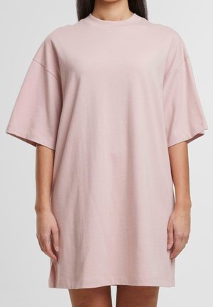 Vrouw draagt een losse, korte mouwen, lichtroze T-shirtjurk tot op de knieën, staand tegen een effen lichte achtergrond.