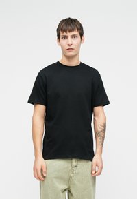 Μαύρη βαμβακερή μπλούζα με κονά μανίκια, στρογγυλή λαιμόκοψη και ίσια γραμμή, φορεμένη με ανοιχτόχρωμο παντελόνι. Λεπτή υφή, χωρίς σχέδια.