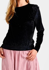 Svart sammetstopp med lång ärm som har en volangkant i halsen, elastisk nederkant och en subtil glans. Kombinationen med en rosa kjol.