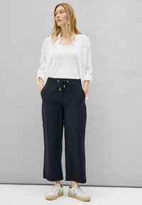 Pantalons larges bleu marine avec taille élastique et cordon de serrage, associés à un débardeur blanc et une chemise blanche ample. Modèle portant des baskets beiges.