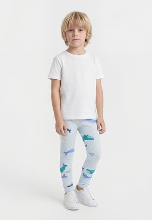 Blond dieťa stojace, oblečené v jednoduchom bielom tričku, svetlomodrých legínach s motívom veľrýb a bielych teniskách na jednofarebnom bielom pozadí.