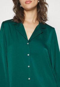Blouse en soie vert émeraude avec col pointu et manches longues, dotée d'une rangée de cinq boutons en métal argenté sur le devant. Texture lisse.