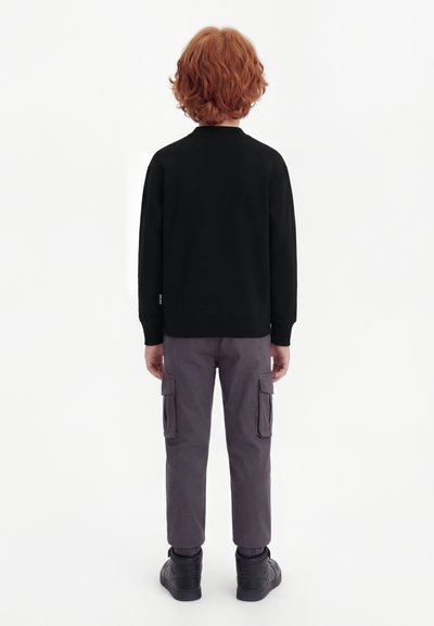 Παιδί με σγουρά κόκκινα μαλλιά στέκεται με την πλάτη, φοράει μαύρο πουκάμισο μακρυμάνικο, γκρι παντελόνι cargo και μαύρα αθλητικά παπούτσια ψηλής κορυφής.