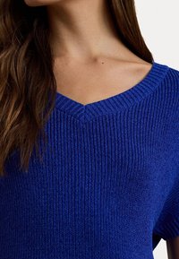 Modrý pletený svetr s výstřihem do V, s žebrovanou texturou a krátkými rukávy. Detailní záběr ukazuje jemné detaily látky a vzor stehů.