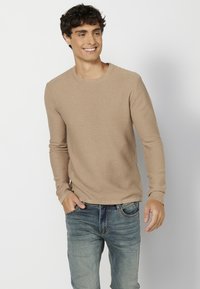 Khaki textúrájú, hosszú ujjú póló, kerek nyakkivágással, világoskék farmernadrággal párosítva, kezével a zsebében, világos háttérrel.