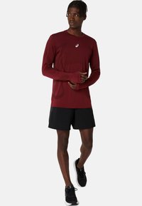 Bordeauxrotes langärmeliges Shirt mit Rundhalsausschnitt und Daumenlöchern, kombiniert mit schwarzen Shorts und schwarzen Sportschuhen, in neutraler Pose stehend.