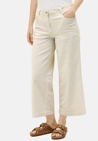 Beige weite Cord-Hosen mit vertikalen Streifen, ausgestattet mit Seitentaschen und einem Knopfverschluss, kombiniert mit braunen Sandalen.