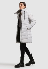 Femeie îmbrăcată cu o haină puffer lungă de culoare gri deschis, cu guler înalt, pantaloni leggings negri și cizme negre până la gleznă, pe un fundal simplu.
