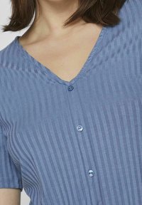 Žena v modrém žebrovaném tričku s krátkým rukávem a V-výstřihem, které má uprostřed tři knoflíky.