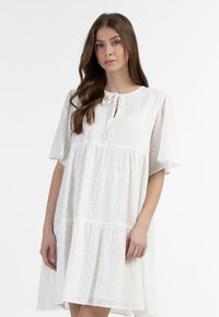 Hvid kjole med korte puffærmer, bindebånd i halsen og lagdelt design. Stoffet har et tekstureret prikkemønster og en løs pasform.