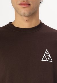 Camiseta de algodón marrón con cuello redondo y un logo triangular blanco en el lado izquierdo del pecho que presenta las letras H, U y F.