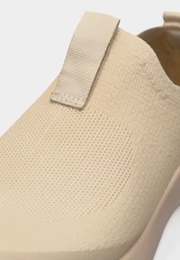 Κοντινό πλάνο μπεζ παπουτσιού χωρίς κορδόνια με υφασμάτινη υφή και υφασμάτινη θηλιά στο πίσω μέρος, τοποθετημένο σε λευκό φόντο.