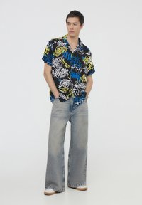 Camicetta nera a maniche corte con motivi colorati di graffiti, abbinata a jeans ampi di color blu chiaro e sneakers beige.