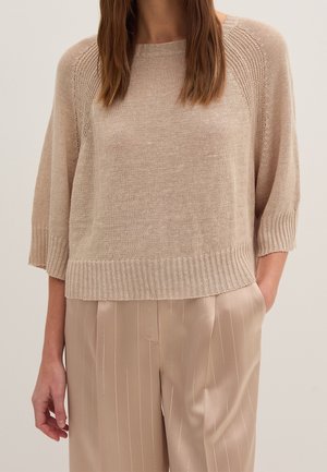 Woman wearing a beige knit sweater with three-quarter sleeves and beige satin pinstriped pants, with one hand in pocket.