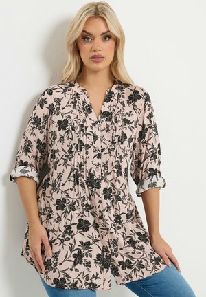 Kvinde med blondt hår iført en lyserød skjorte med sort blomstermønster og opkruede ærmer samt blå jeans mod en hvid baggrund.