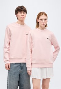 Νεαρός άνδρας και γυναίκα που φορούν ταιριαστές ανοιχτό ροζ φούτερ, στέκονται δίπλα-δίπλα μπροστά σε απλό λευκό φόντο.