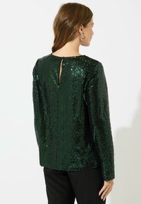 Top verde con paillettes, maniche lunghe, scollatura rotonda e dettaglio a chiave sulla schiena. Presenta una superficie texturizzata e lucida. Indossato con pantaloni neri.
