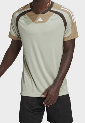 Mężczyzna ubrany w beżowo-brązową sportową koszulkę Adidas i czarne szorty, stojący na jednolitym tle.