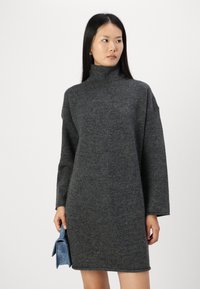 Grå strikket høyhalset kjole med lange ermer og rett snitt. Stoffet ser mykt og teksturert ut. En blå veske holdes i høyre hånd.