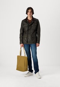 Veste cirée vert foncé avec plusieurs poches, col marron et boutons dorés. Associée avec un jean bleu et un sac fourre-tout beige. Baskets blanches.