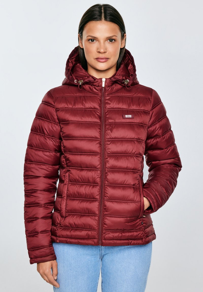 Žena v vínově červené prošívané bundě s kapucí a zapínáním na zip, s jednou rukou v kapse, v kombinaci se světle modrými džíny na jednobarevném pozadí.