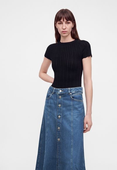 Femme aux cheveux bruns lisses portant un haut noir à manches courtes et une jupe en jean bleue à boutons sur le devant, debout devant un fond uni.