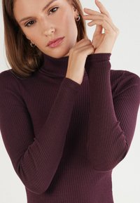 Burgunderfarbenes, geripptes Rollkragenoberteil mit langen Ärmeln, das eine figurbetonte Silhouette und eine weiche Textur bietet. Verlängerte Ärmel betonen das Design.