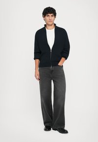 Mörkblå ribbad zip-up cardigan med korta ärmar, bärs över en vit skjorta, i kombination med svarta vida byxor och svarta skor.