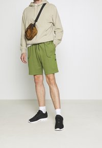 Krémová mikina s kapucí, zelené šortky s předními kapsami, černé tenisky a bílé ponožky. Model pózuje na jednobarevném pozadí.