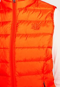 Orange puffy väst gjord av slät, glänsande tyg, med en vertikal dragkedja, horisontell sömnad och en broderad logotyp i röd.