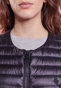 Puffer vest in dark navy, featuring horizontal quilted design, zip closure, and small logo embroidery on the left chest. Gray shirt beneath.