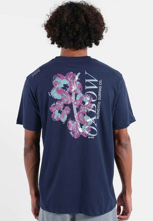 Homme portant un T-shirt bleu marine avec des fleurs d'hibiscus roses et bleues et le texte "OZABLOW Authentic Surfing Co." dans le dos.